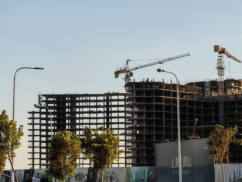 Commercial building under construction with exposed steel framework and cranes against a blue sky